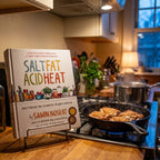Salt, Fat, Acid, Heat on a cookbook stand beside a cast-iron skillet